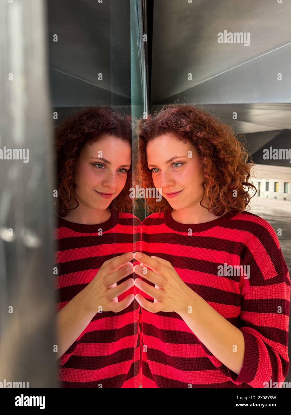 Una donna rossa con un pullover a righe rosse e nere si staglia su una superficie riflettente creando un'illusione simmetrica Foto Stock