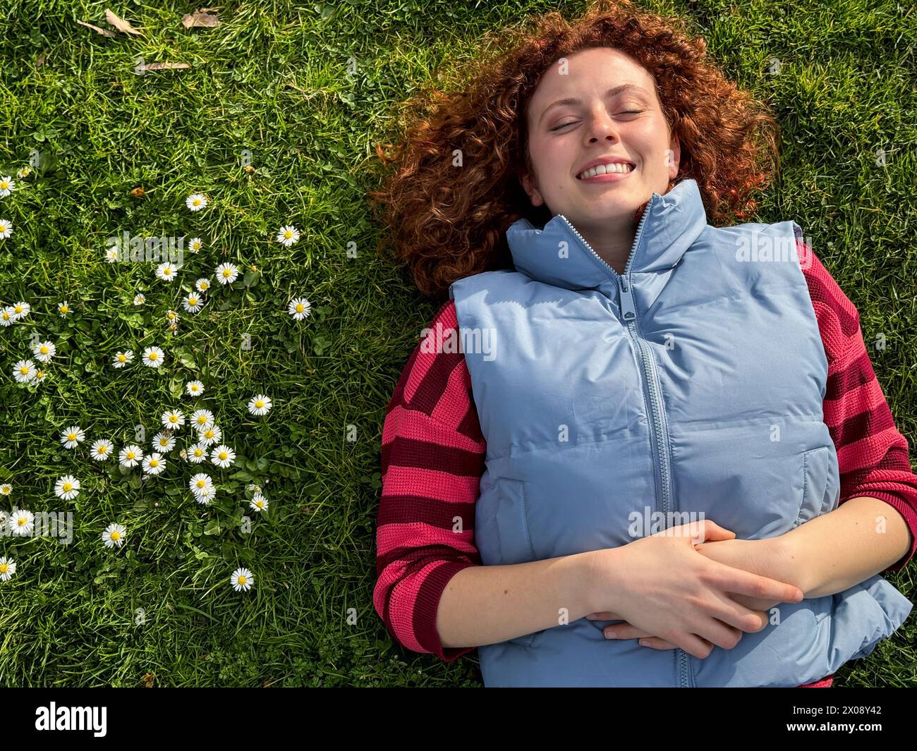 Una donna rossa gioiosa giace su un campo erboso tra margherite, incarnando relax e felicità Foto Stock