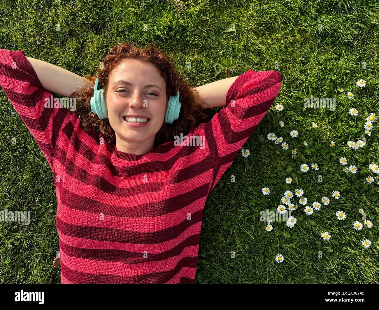 Una giovane e allegra rossa giace sull'erba con le braccia allungate, ascoltando la musica con le sue cuffie wireless, circondate da margherite Foto Stock