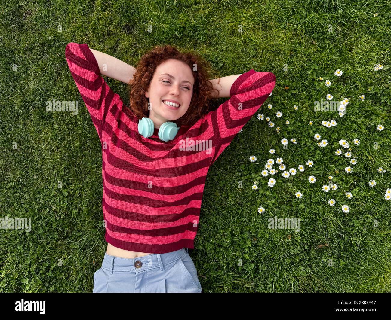 Una giovane donna rossa felice con i capelli rossi ricci giace su un lussureggiante prato verde, circondato da margherite bianche, godendosi una piacevole giornata Foto Stock