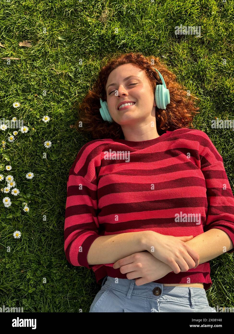 Una giovane donna rossa gode di un momento di pace sdraiata sull'erba con le cuffie, circondata da fiori selvatici Foto Stock