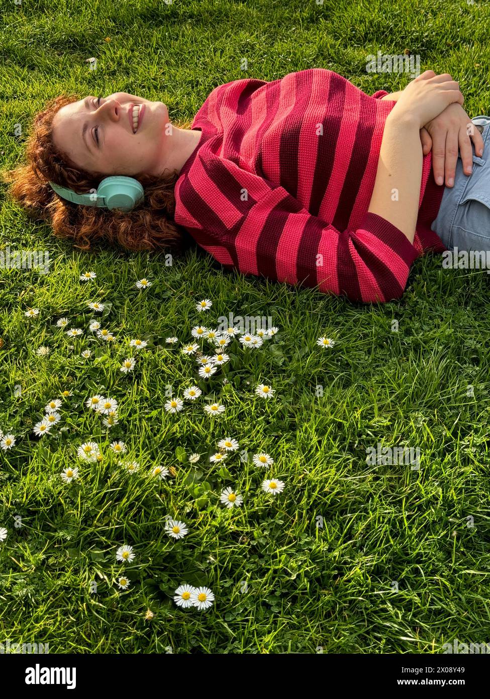 Una giovane donna rossa gioiosa giace su un campo erboso con margherite, indossando le cuffie e godendosi la calda luce del sole Foto Stock