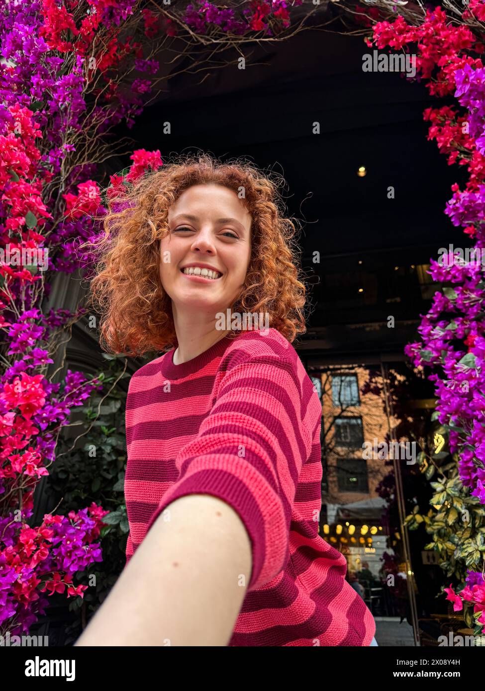 Allegra giovane donna rossa con capelli ricci sorridenti sotto un vivace arco floreale in un ambiente urbano Foto Stock