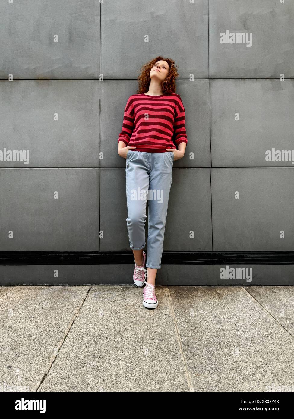 Una giovane donna rossa dai capelli ricci con un pullover a righe e pantaloni grigi si appoggia a una parete grigia testurizzata, guardando con attenzione Foto Stock