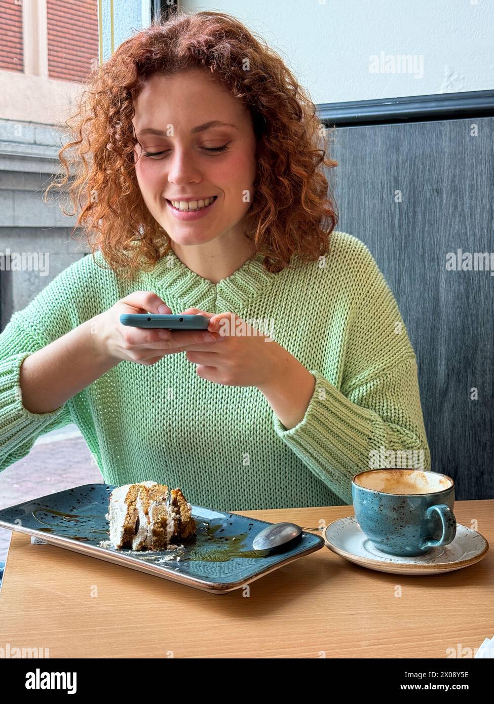 Una donna rossa gioiosa si siede al tavolo di un bar e scatta foto con il suo smartphone, con una fetta di torta parzialmente mangiata e una tazza di caffè Foto Stock