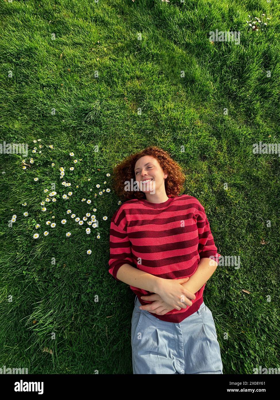Una donna allegra rossa giace su un'erba verde brillante, il suo sorriso irradia gioia tra le piccole margherite bianche Foto Stock