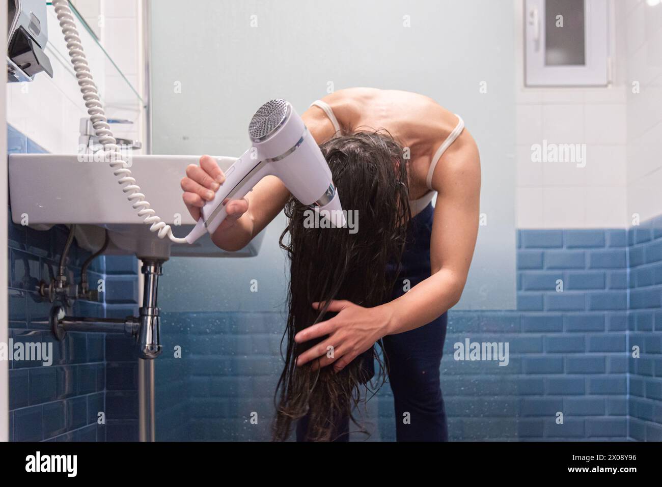 Una donna in bagno si piega in avanti per asciugarsi i capelli bagnati con un asciugacapelli bianco dopo una doccia Foto Stock
