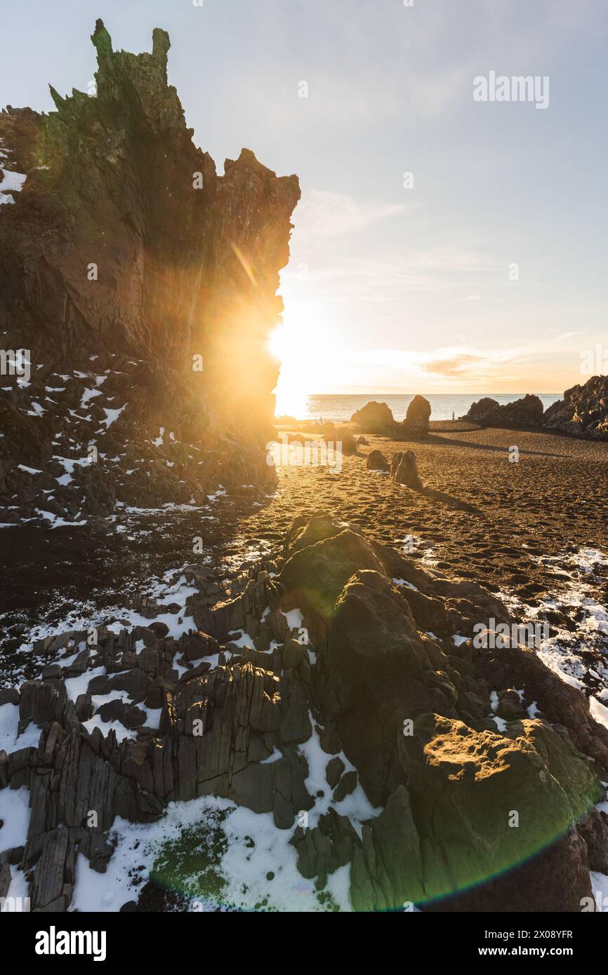 Il sole brilla intorno a un'aspra scogliera basaltica su una spiaggia islandese, evidenziando le impressionanti formazioni geologiche e il delicato tocco della neve Foto Stock