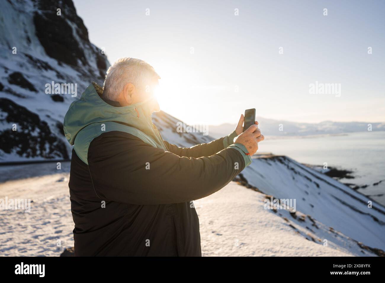 Un uomo cattura il momento al telefono, con il sole invernale che proietta un bagliore radioso sulla costa innevata dell'Islanda Foto Stock