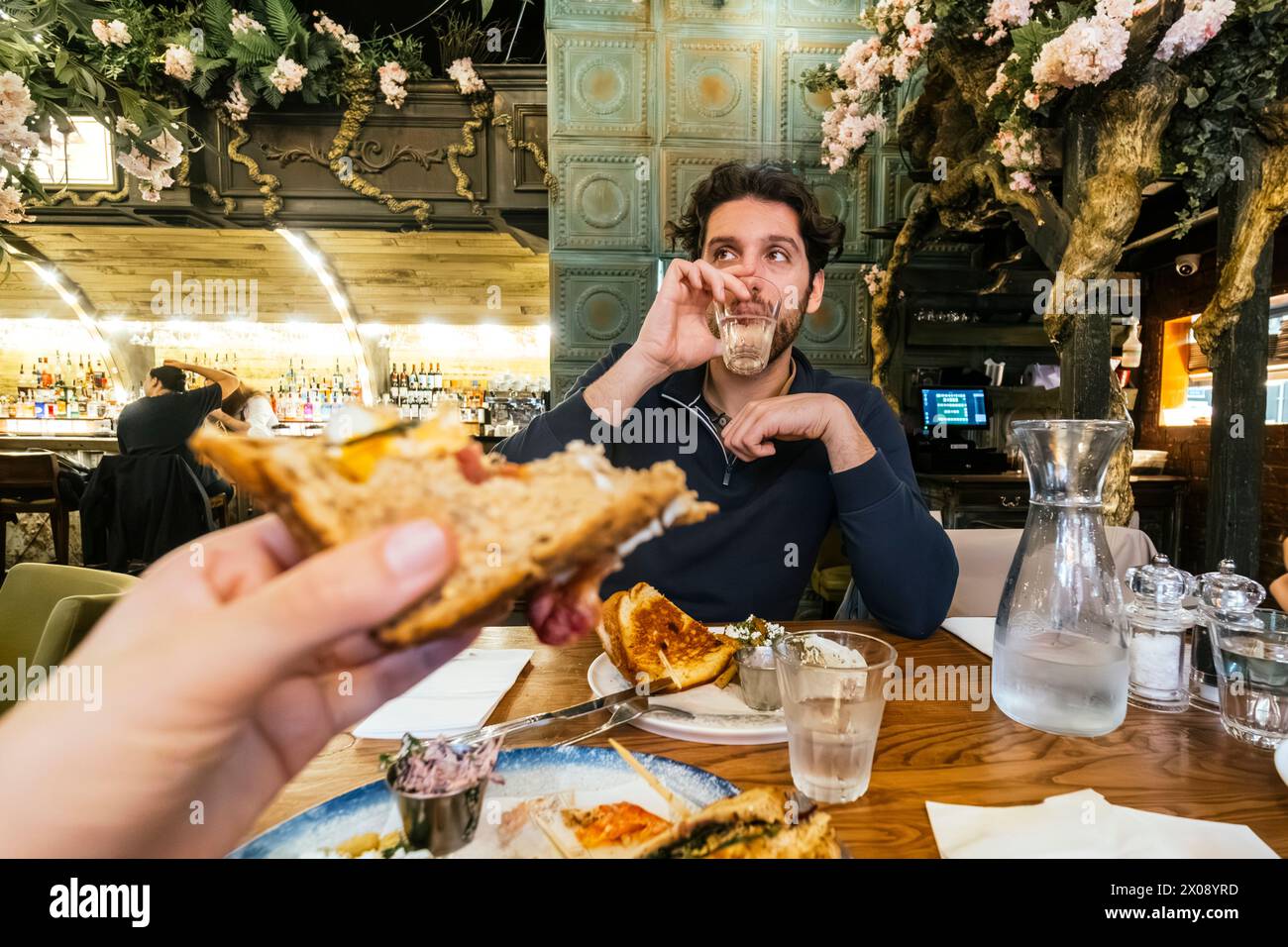 Un uomo prende un piacevole boccone del suo panino, fermandosi per assaporarne il sapore, nell'incantevole atmosfera di un bar con decorazioni botaniche Foto Stock