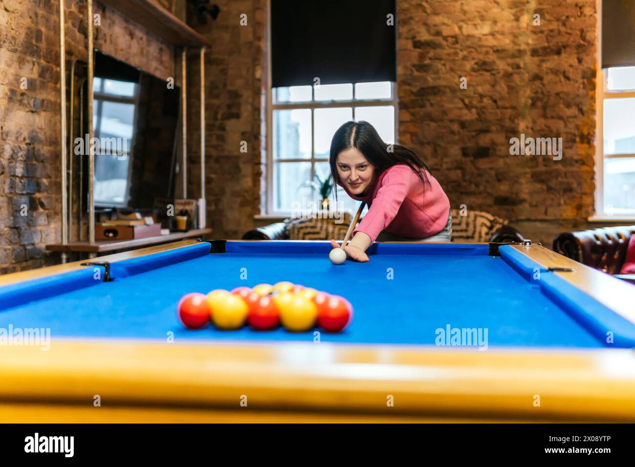Una giovane donna con un'espressione concentrata che gioca a biliardo in un ambiente interno accogliente, potenzialmente con gli amici fuori dalla cornice Foto Stock