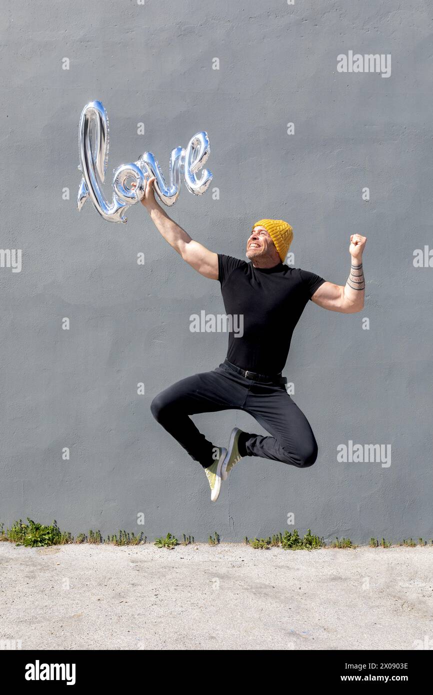 Un uomo che brilla in un abbigliamento casual che salta con gioia mentre tiene in mano un palloncino d'amore lucente. Foto Stock