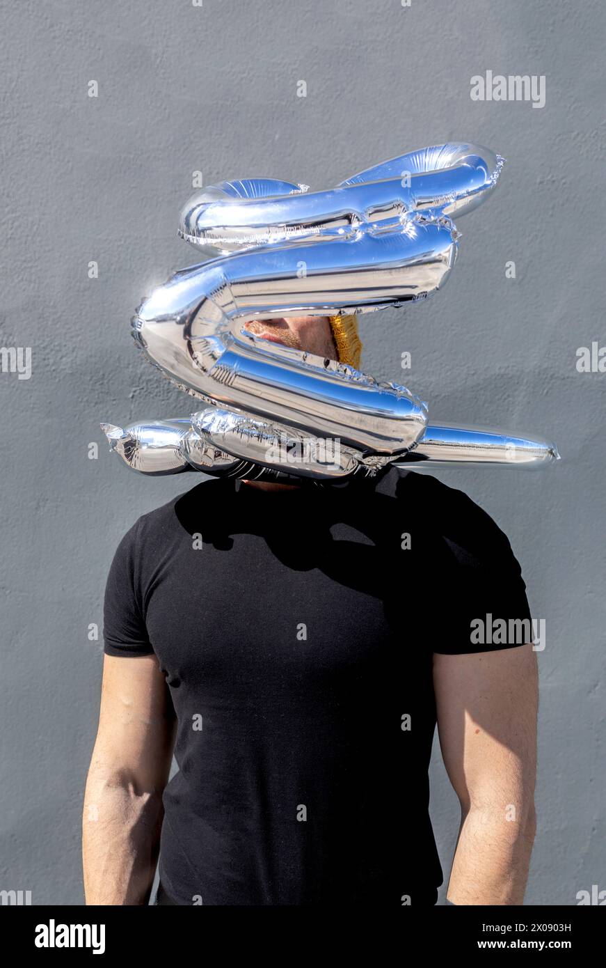 Un uomo irriconoscibile si staglia su uno sfondo grigio con palloncini d'argento che scrivono l'amore fluttuando sopra la testa, nascondendo il suo volto. Foto Stock