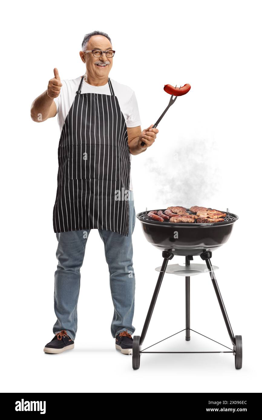 Uomo maturo che cuoce la carne su un barbecue e tiene in mano una salsiccia con una forchetta isolata su sfondo bianco Foto Stock