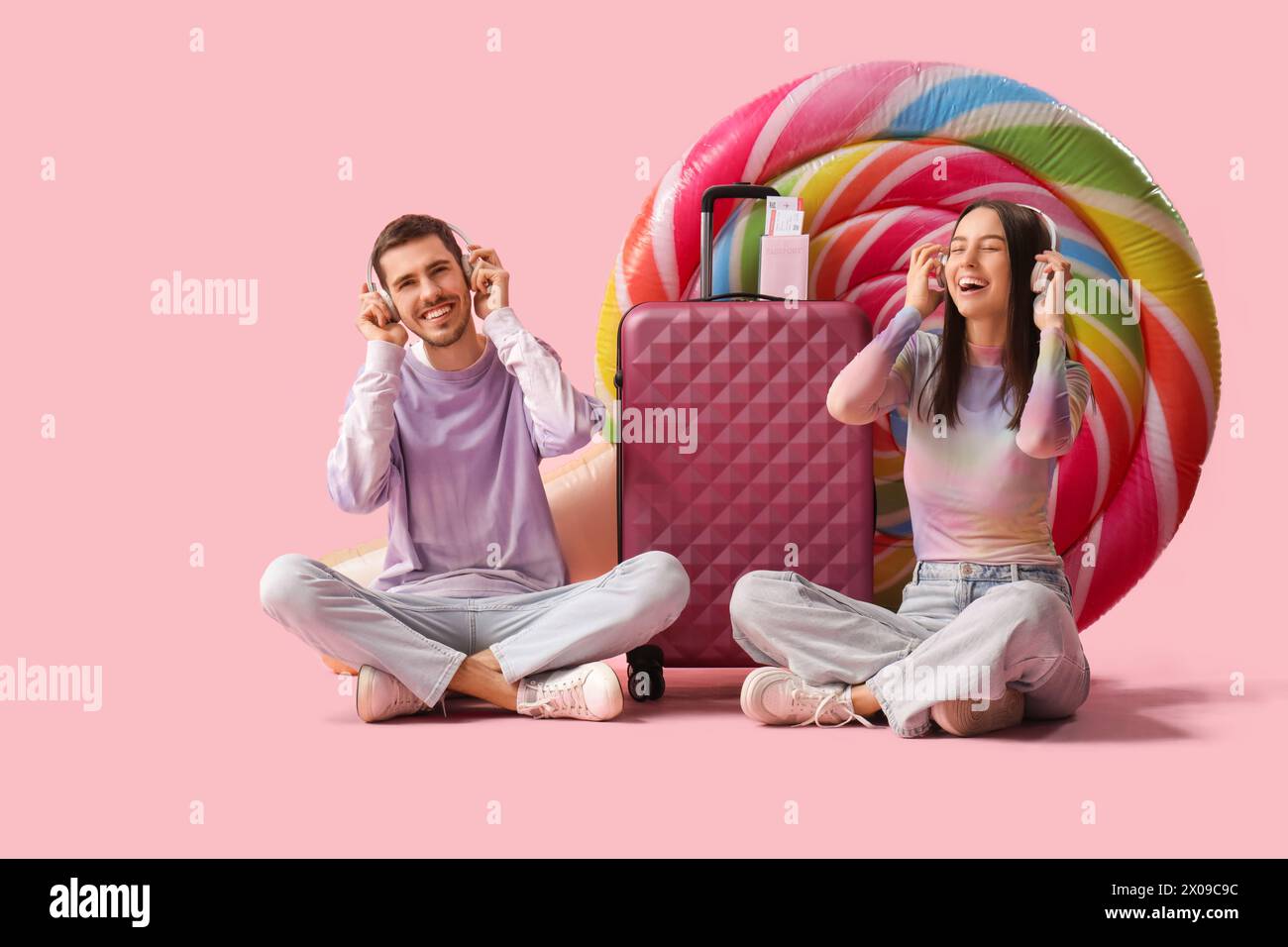 Bella giovane coppia felice di turisti in cuffie con valigia, biglietti e materasso gonfiabile seduti su sfondo rosa Foto Stock