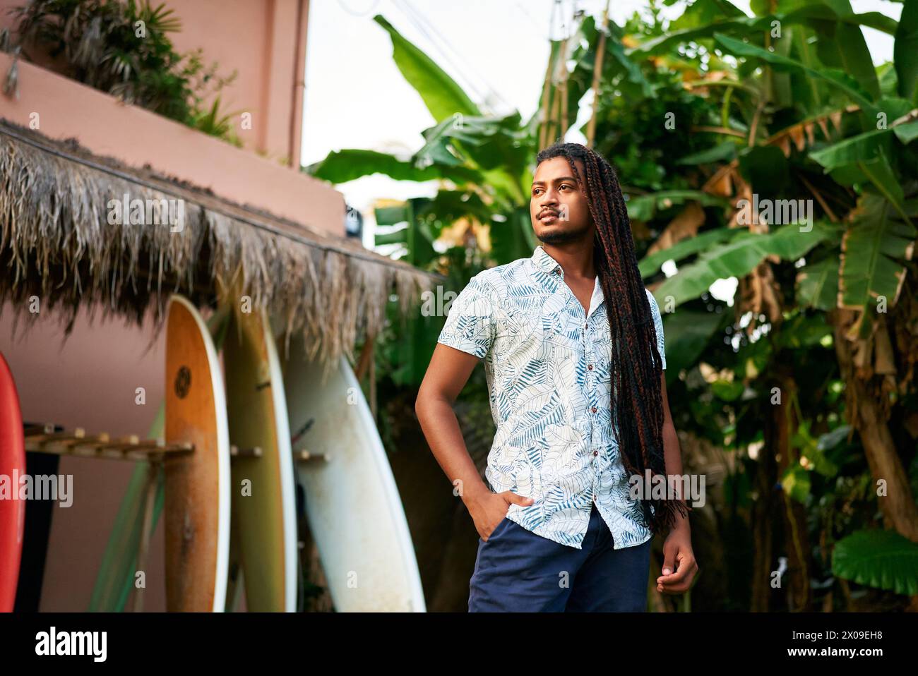 Istruttore di surf con la pelle scura si erge tra tavole colorate in un ambiente tropicale. Il coach BIPOC attende gli studenti per una lezione di piante verdi lussureggianti Foto Stock