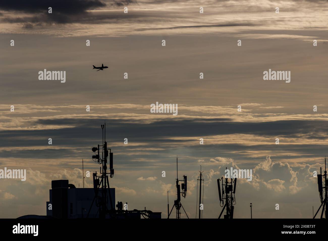 Paesaggio cittadino con un aereo commerciale che vola nel cielo del tramonto, tra gli edifici e le antenne Foto Stock