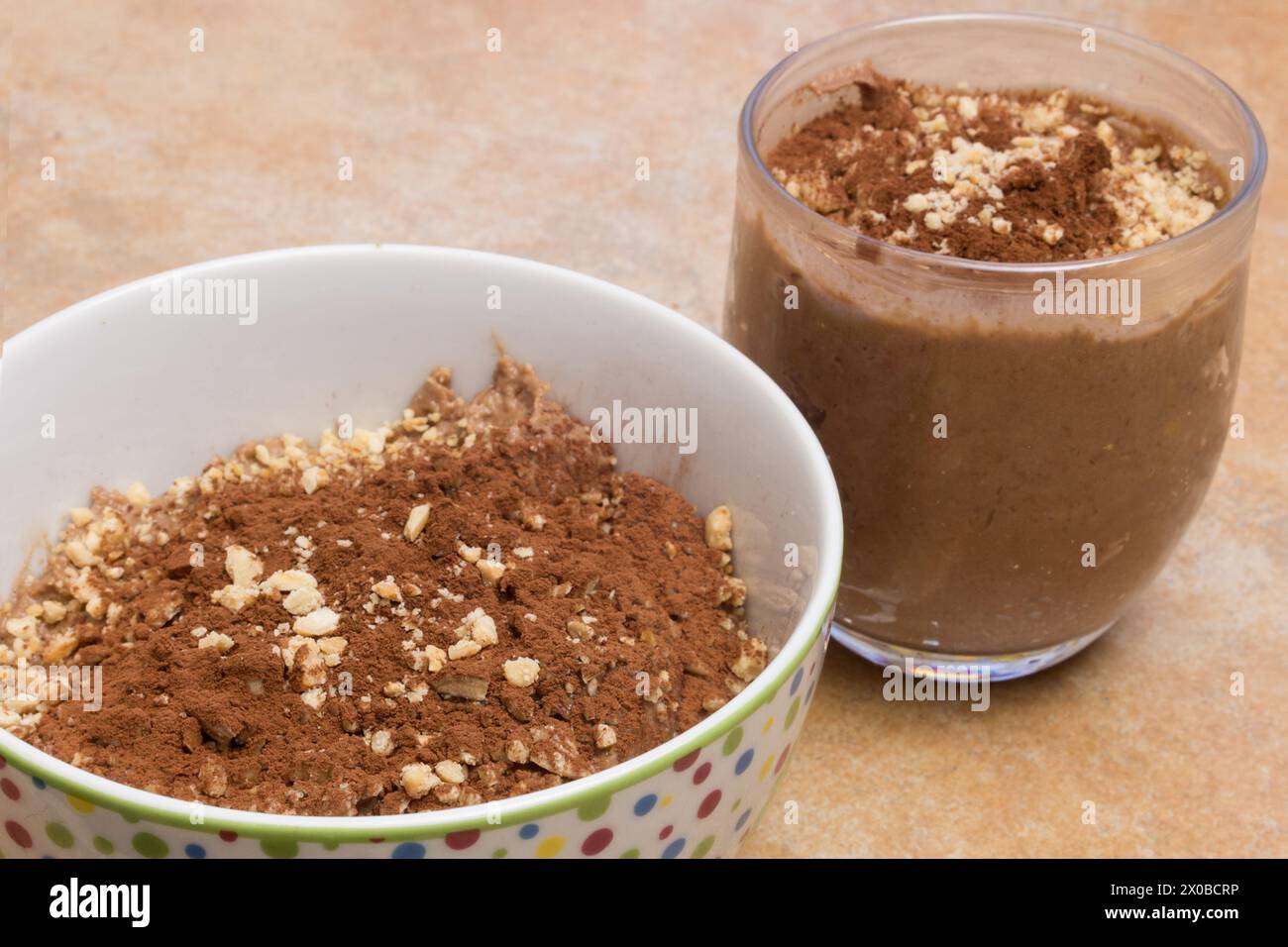 Primo piano di un bicchiere e di una ciotola ripiena di mousse al cioccolato arricchita con cacao e mandorle su un piano di lavoro. Dessert facili e veloci. Foto Stock
