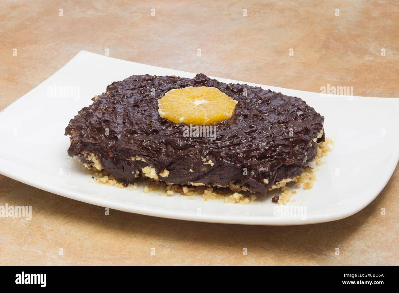 Un ricco dessert al cioccolato condito con una vivace fetta di arancia fresca, appoggiato su un piatto bianco contro un ripiano beige, che mette in risalto il dessert Foto Stock