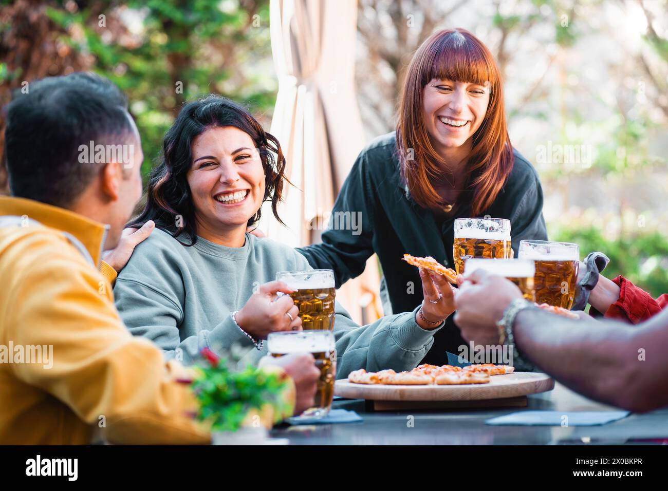 Gli amici più stretti condividono un'abbondante risata con birra e pizza ghiacciate in un giardino all'aperto - un momento di gioia e cameratismo - il festival delle attività del fine settimana Foto Stock