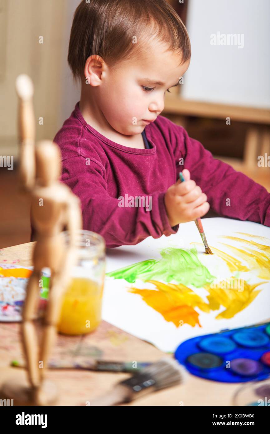 Scatto verticale di un bambino piccolo che dipinge con colori acrilici colorati sulla sua tela bianca sul suo tavolo da lavoro, dove ci sono diversi pennelli, dipinge un Foto Stock