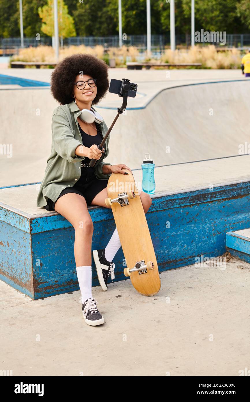 Una giovane donna afroamericana con capelli ricci seduta su una panchina con uno skateboard in un vivace parco di skate. Foto Stock