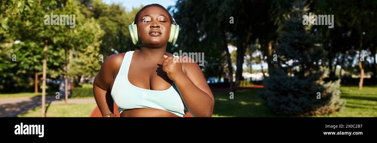 Una donna afroamericana in abbigliamento sportivo, che abbraccia la sua positività corporea, corre in un parco indossando le cuffie, sintonizzandosi sui suoni della natura. Foto Stock