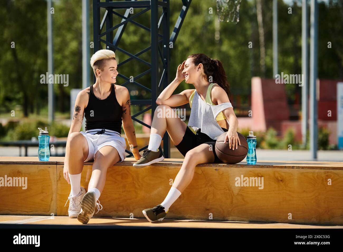 Due giovani donne atletiche si godono una pausa sulla panchina di legno dopo aver giocato a basket all'aperto in estate. Foto Stock