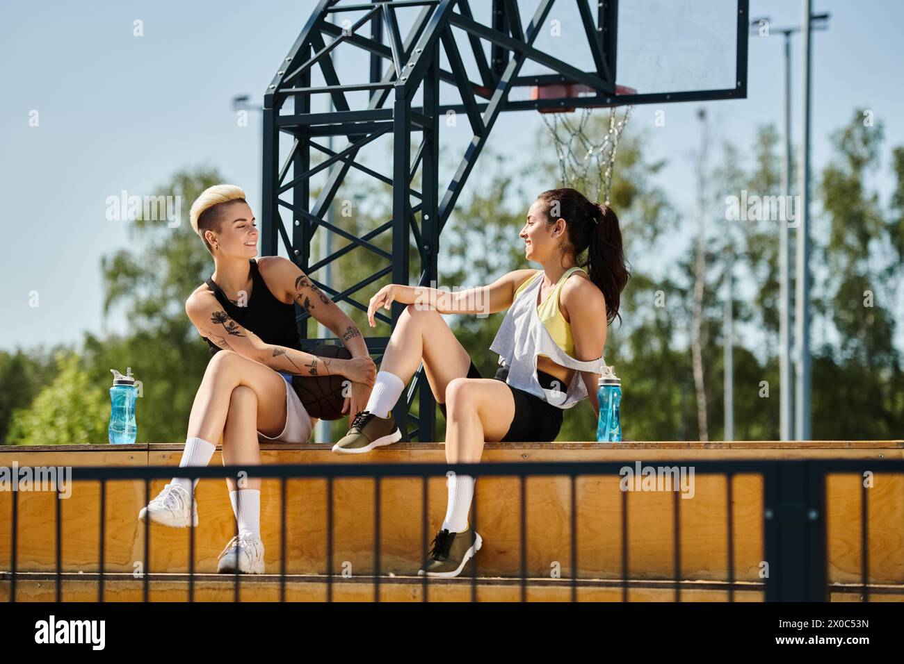 Due giovani donne, atletiche e vibranti, si impegnano in una conversazione su una panchina in un ambiente soleggiato all'aperto, immersi in un momento di legame e risate. Foto Stock