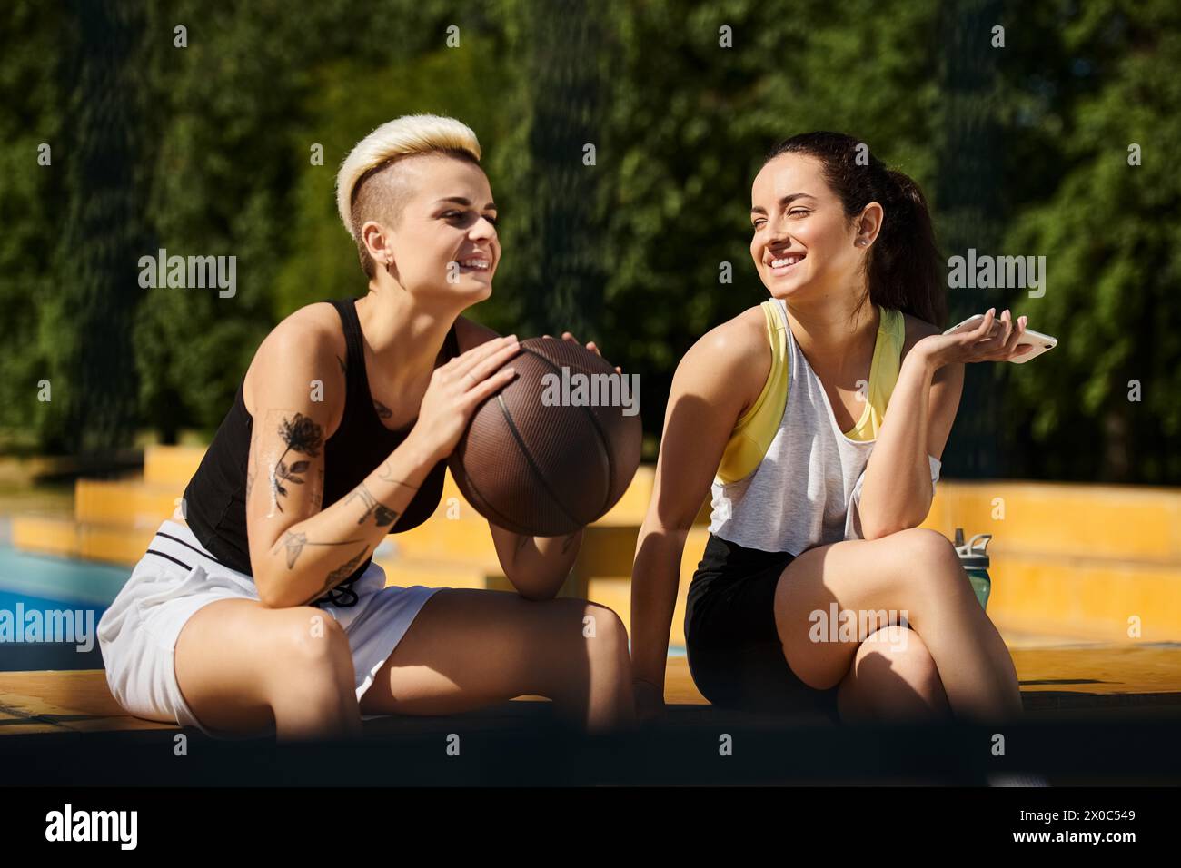 Due giovani donne sedute insieme, che tengono una pallacanestro, mostrano il loro amore per lo sport in una giornata di sole. Foto Stock