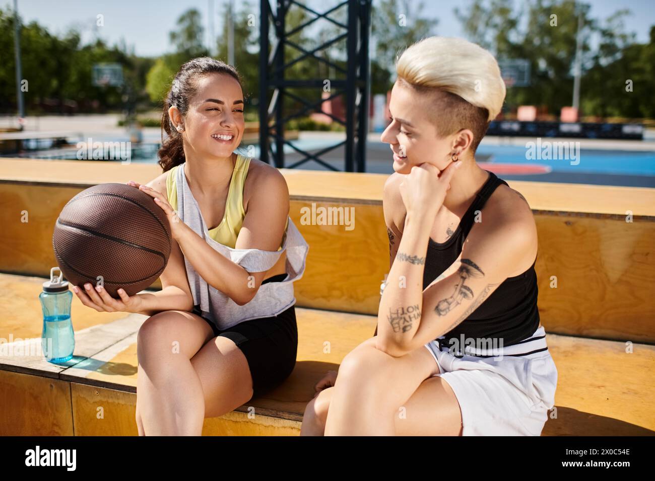 Due donne atletiche sedute fianco a fianco, che tengono una pallacanestro e si godono una giornata estiva all'aperto. Foto Stock
