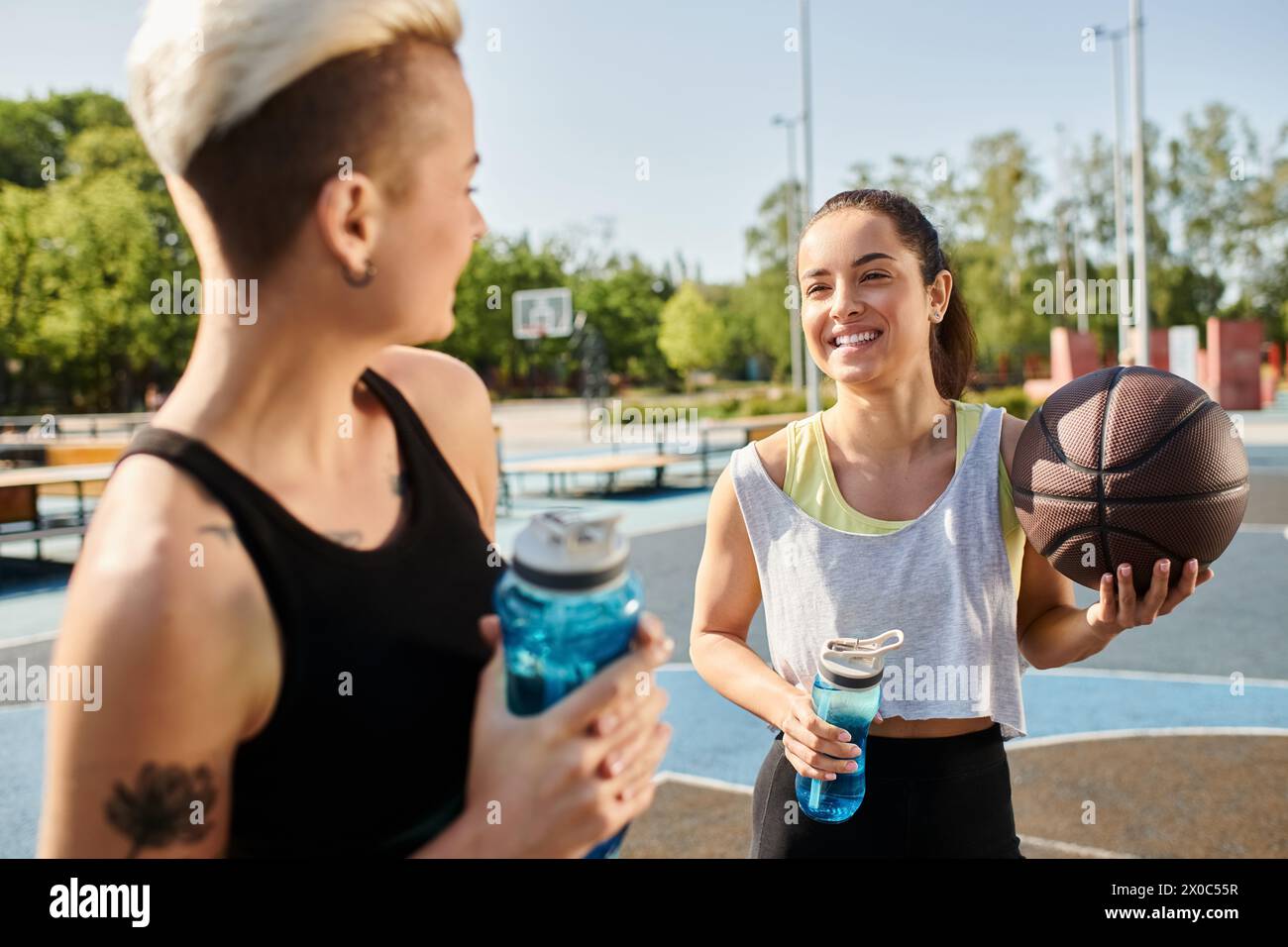 Due giovani donne vestite in estate, in piedi fianco a fianco, che tengono una pallacanestro in un ambiente all'aperto. Foto Stock