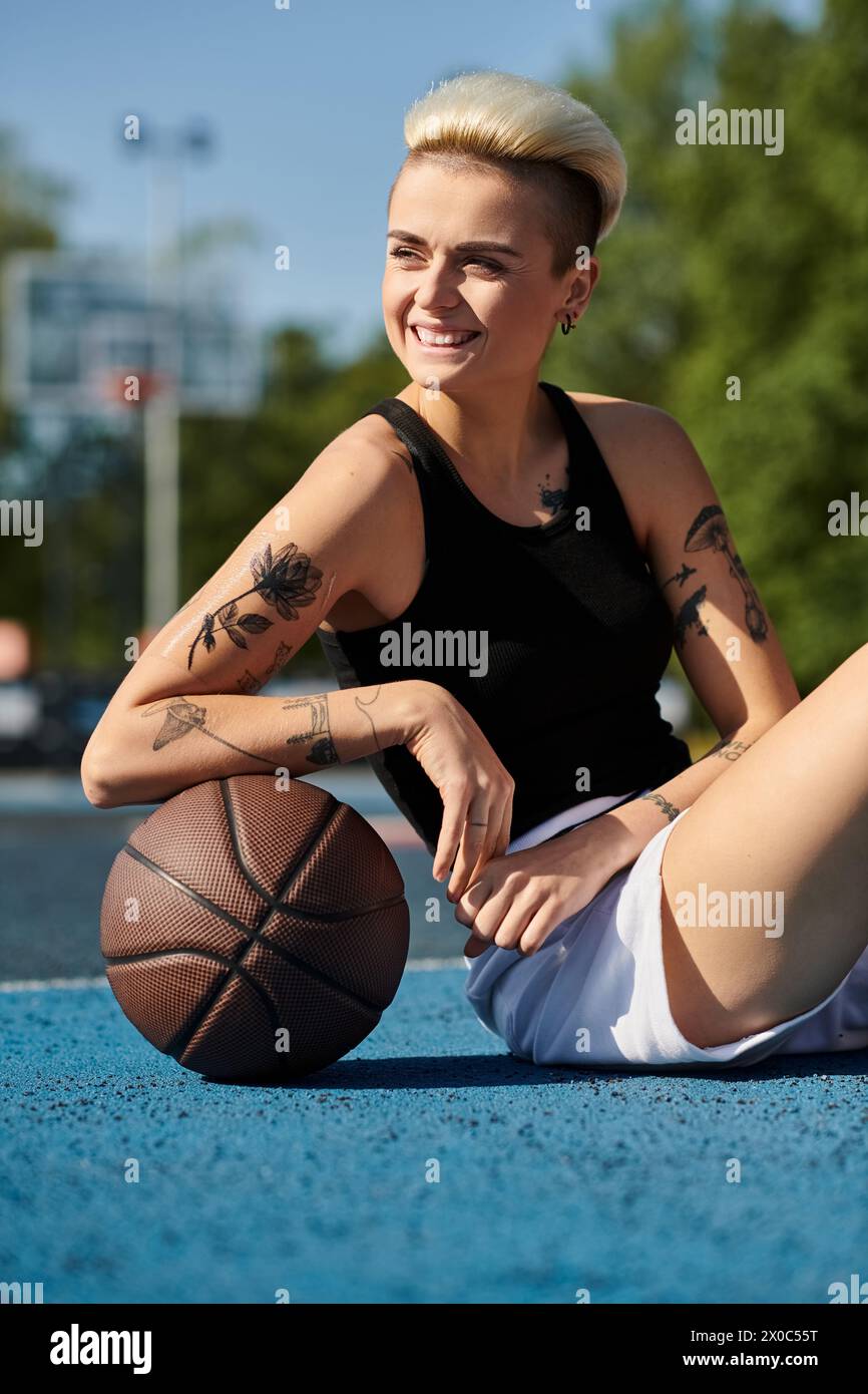 Una giovane donna con i capelli corti e i tatuaggi siede a terra, tenendo una pallacanestro in un ambiente urbano all'aperto. Foto Stock