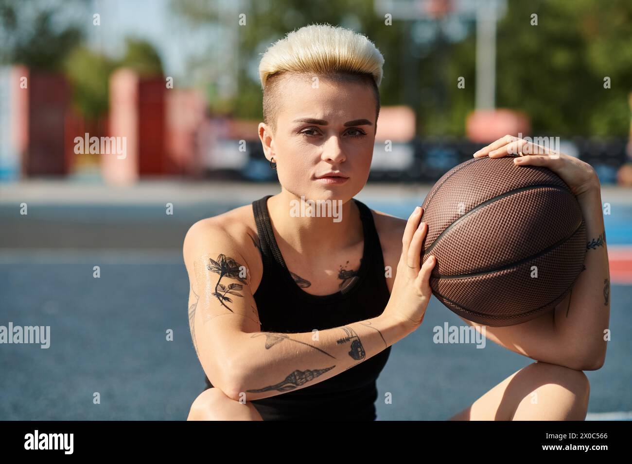 Una giovane donna con i capelli corti e i tatuaggi seduta a terra, che tiene in mano una pallacanestro, persa nel pensiero. Foto Stock