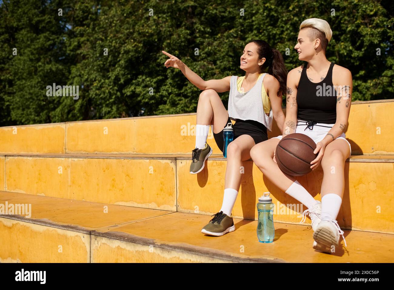 Due giovani donne, amiche atletiche, si siedono a stretto contatto dopo aver giocato a basket all'aperto in una giornata estiva. Foto Stock