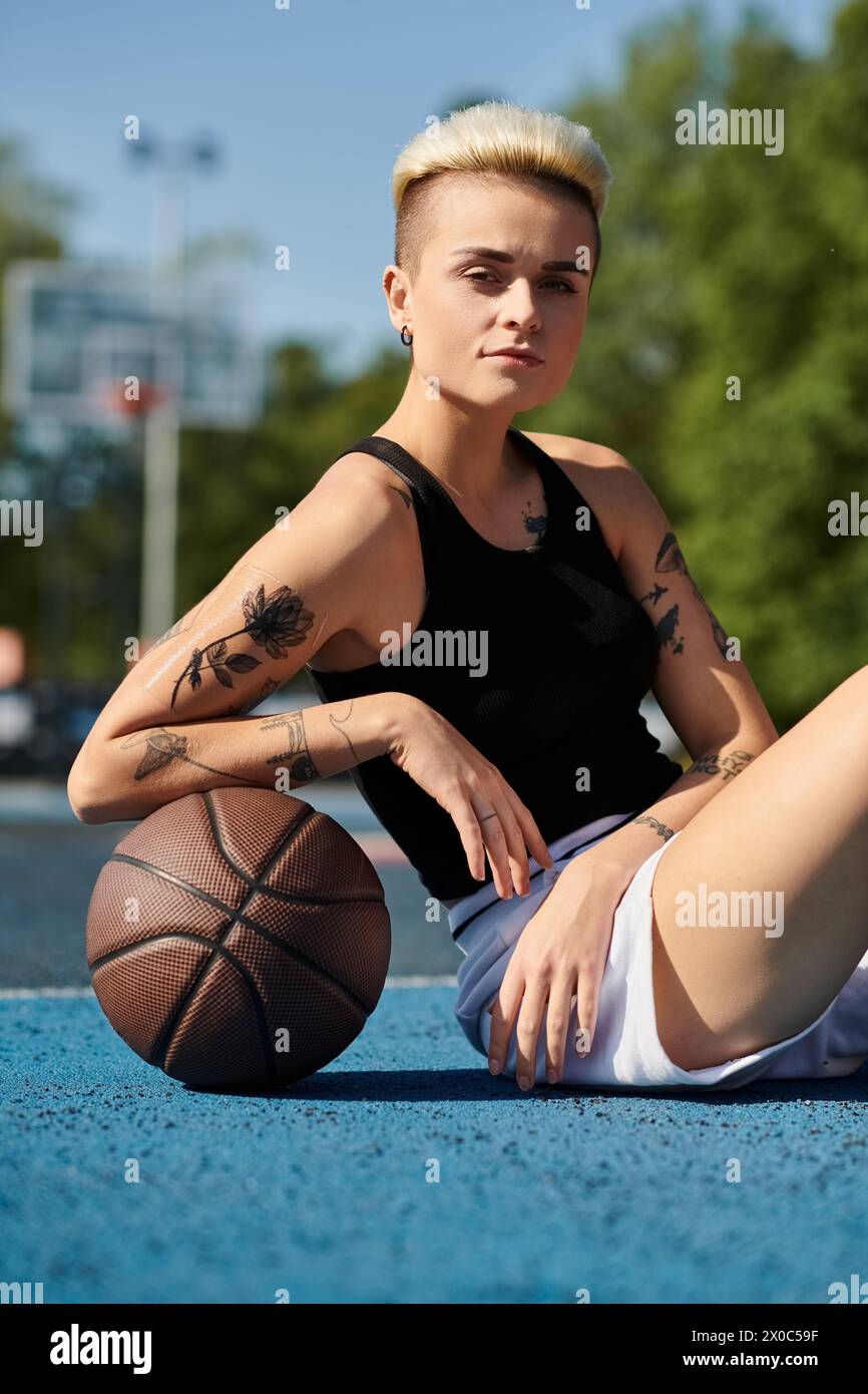 Una giovane donna con i tatuaggi si siede a terra, tenendo in mano una pallacanestro, persa nel pensiero. Foto Stock