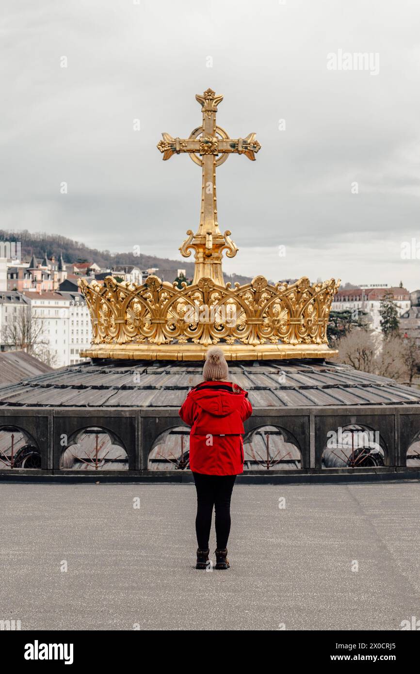 Una ragazza vestita di rosso si erge davanti a un monumento religioso nella città di culto di Lourdes Foto Stock