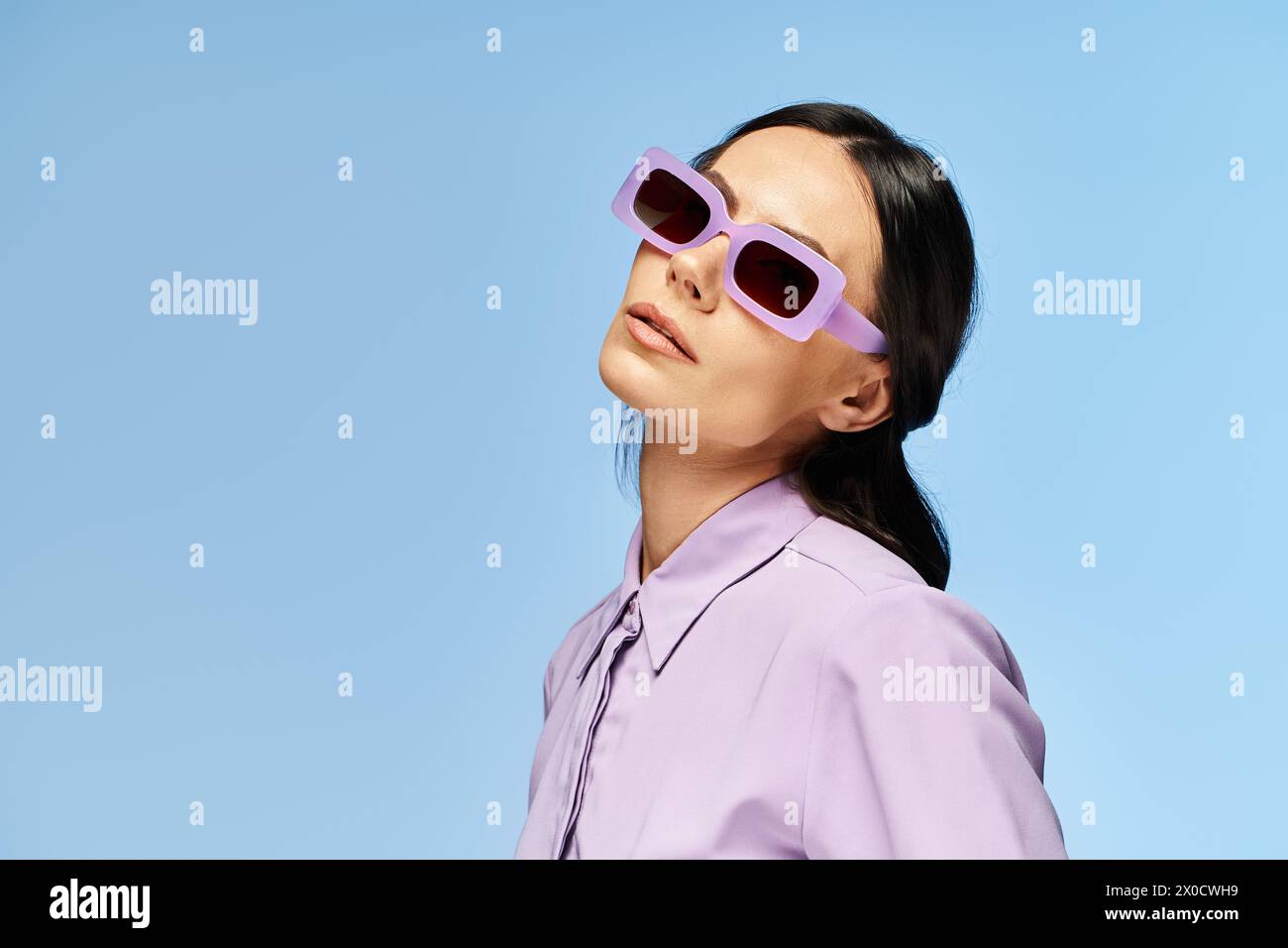 Una donna elegante con occhiali da sole e camicia viola si posa su uno sfondo blu in un ambiente estivo da studio. Foto Stock