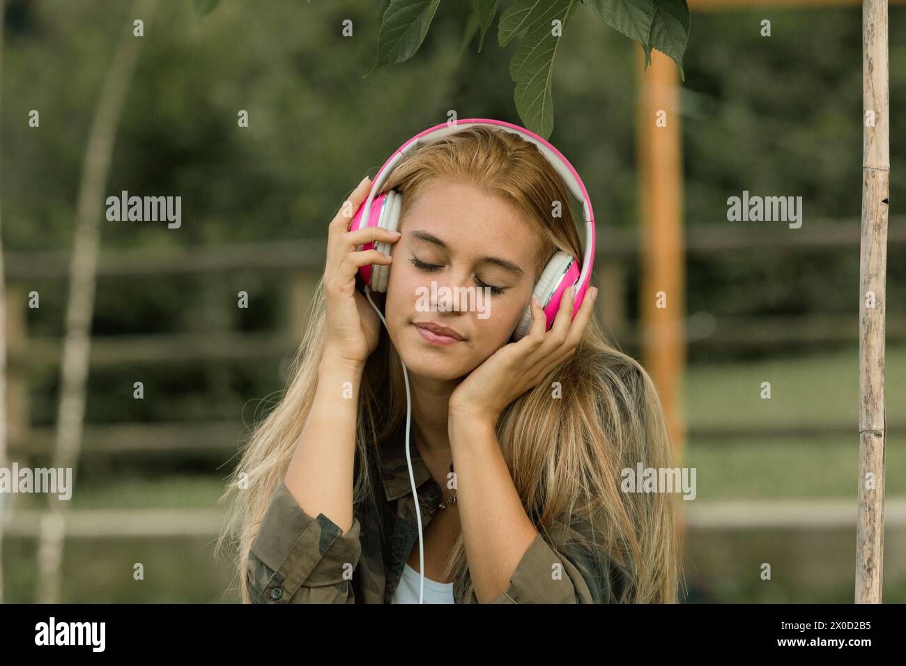 Una giovane donna ama la musica con le cuffie rosa, gli occhi chiusi in un momento sereno, circondati dalla natura Foto Stock