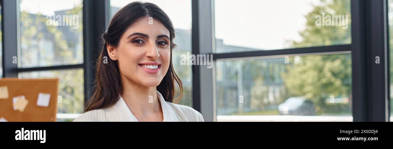 Una donna d'affari sta di fronte a una finestra, sorridendo brillantemente mentre la luce del sole illumina il suo volto, incarnando un senso di empowerment e positività. Foto Stock