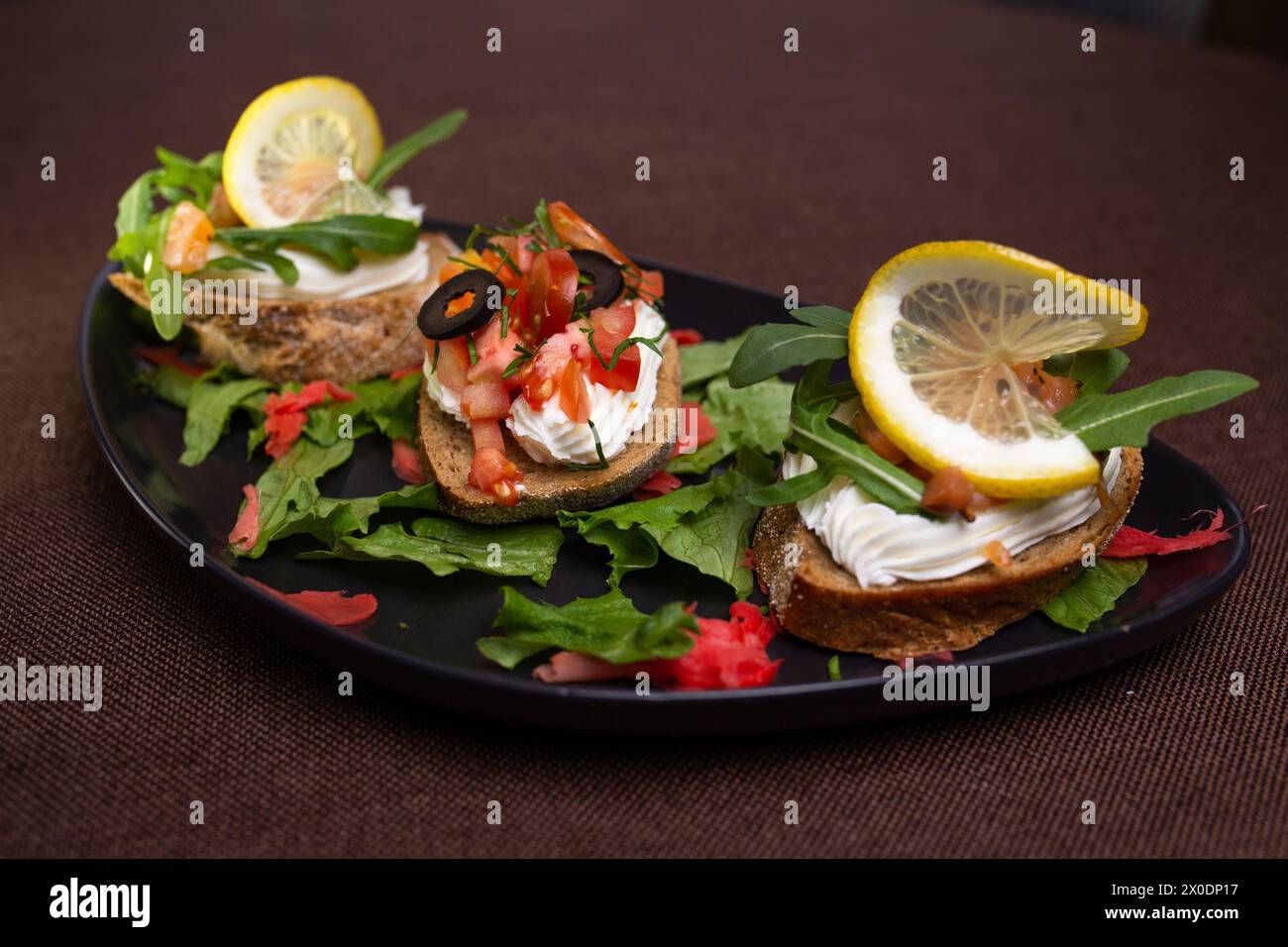 Una varietà di bruschette con formaggio spalmabile, pomodori, olive, limone su un piatto nero, con lattuga e contorno di zenzero sottaceto rosso Foto Stock
