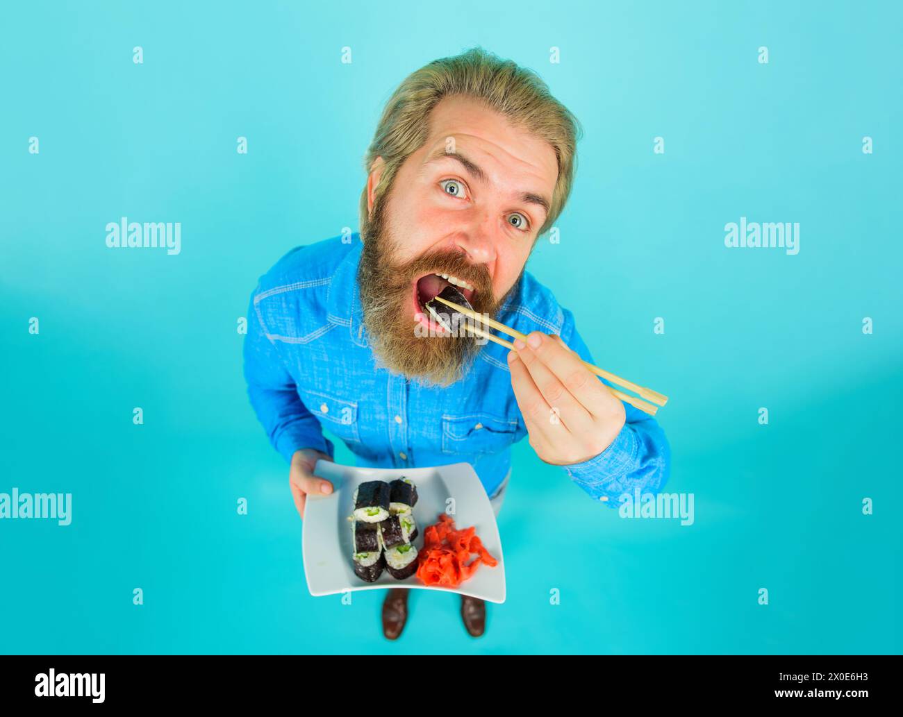 Uomo con barba in camicia casual in denim che mangia sushi roll con bacchette. Consegna di sushi. Cucina tradizionale giapponese. Bell'uomo con piatto di deliziosi Foto Stock