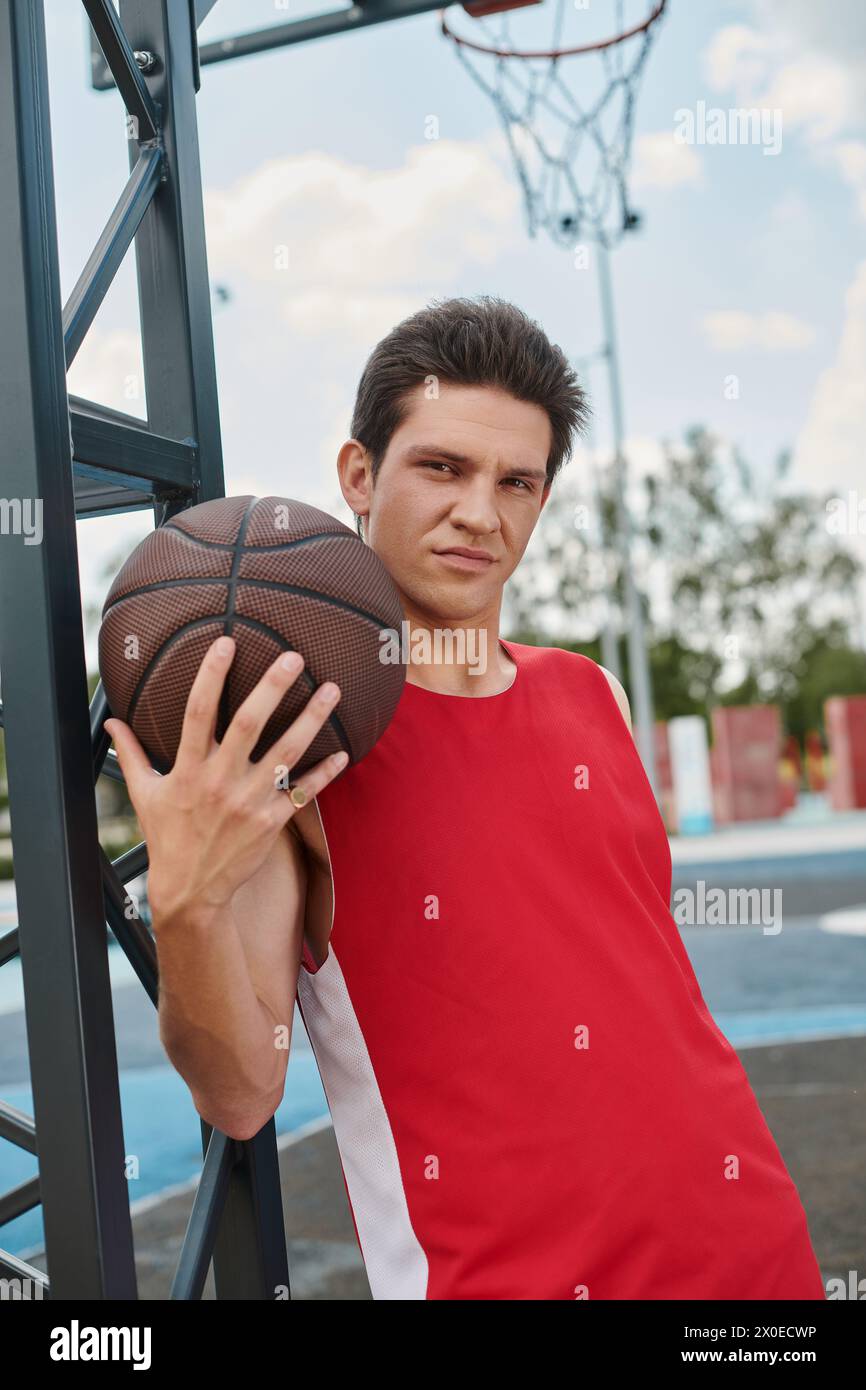 Un giovane con una maglietta rossa regge sapientemente un pallacanestro, pronto a giocare all'aperto in una giornata estiva. Foto Stock