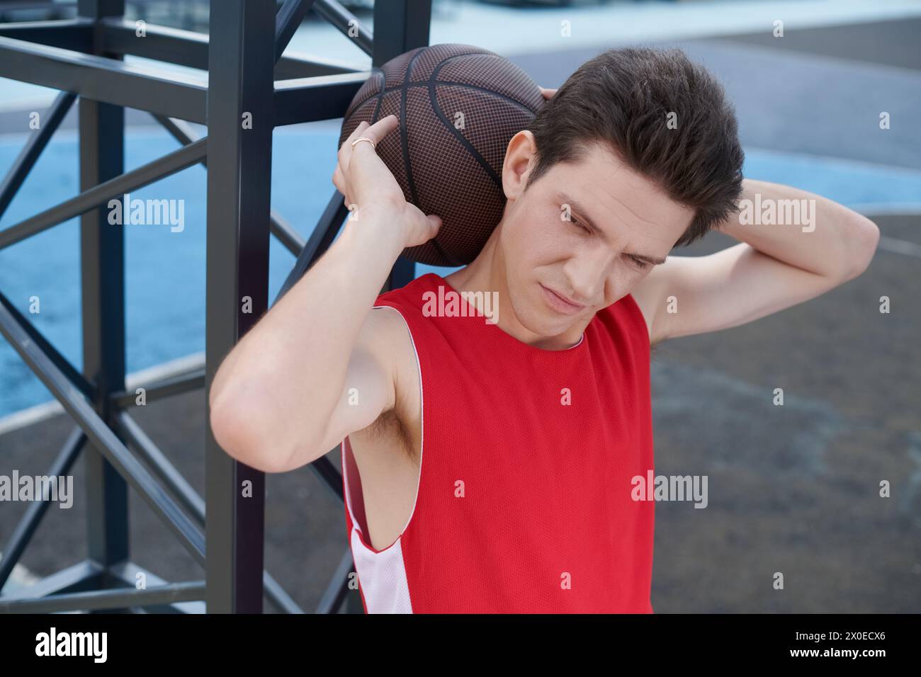 Un giovane con una camicia rossa regge sapientemente un pallacanestro mentre si prepara a giocare all'aperto in una giornata estiva di sole. Foto Stock