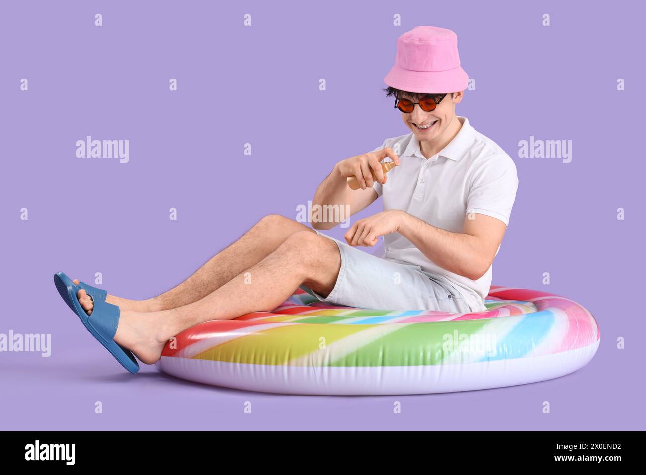 Bel giovane uomo felice con materasso gonfiabile a forma di lecca lecca che applica crema solare su sfondo viola Foto Stock