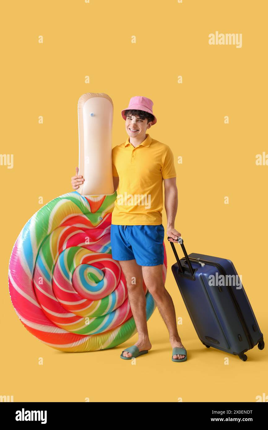Bel giovane uomo felice con materasso gonfiabile a forma di lecca lecca e valigia su sfondo giallo Foto Stock