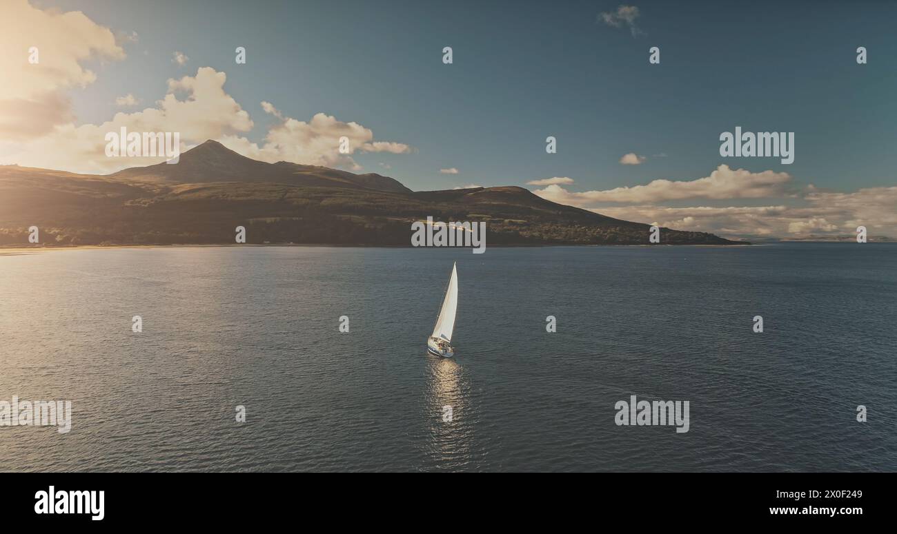 Riflesso del sole di uno yacht a vela sull'antenna della baia dell'oceano. Sole epico d'estate. Crociera di lusso in barca a vela in mare. Isola di montagna di Arran, Scozia con foresta verde sulla costa. Morbide nuvole sopra il supporto Foto Stock