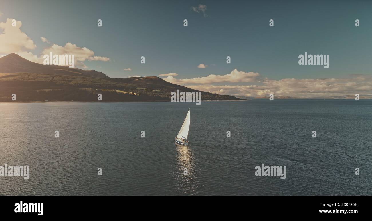 Riflesso del sole di uno yacht a vela sull'antenna della baia dell'oceano. Sole epico d'estate. Crociera di lusso in barca a vela in mare. Isola di montagna di Arran, Scozia con foresta verde sulla costa. Morbide nuvole sopra il supporto Foto Stock