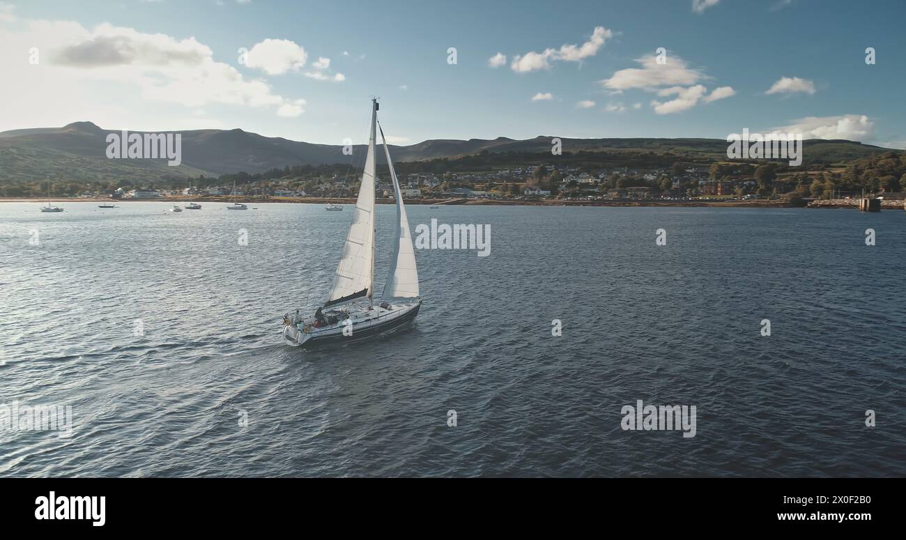 Città portuale con terminal merci e yacht sull'oceano al porto di Brodick. Barca a vela nel golfo del mare. Crociera a bordo di una nave passeggeri che corre a baia con la costa dell'isola di Highlang. Il sole si riflette sulla barca a vela. Paesaggio marino epico Foto Stock