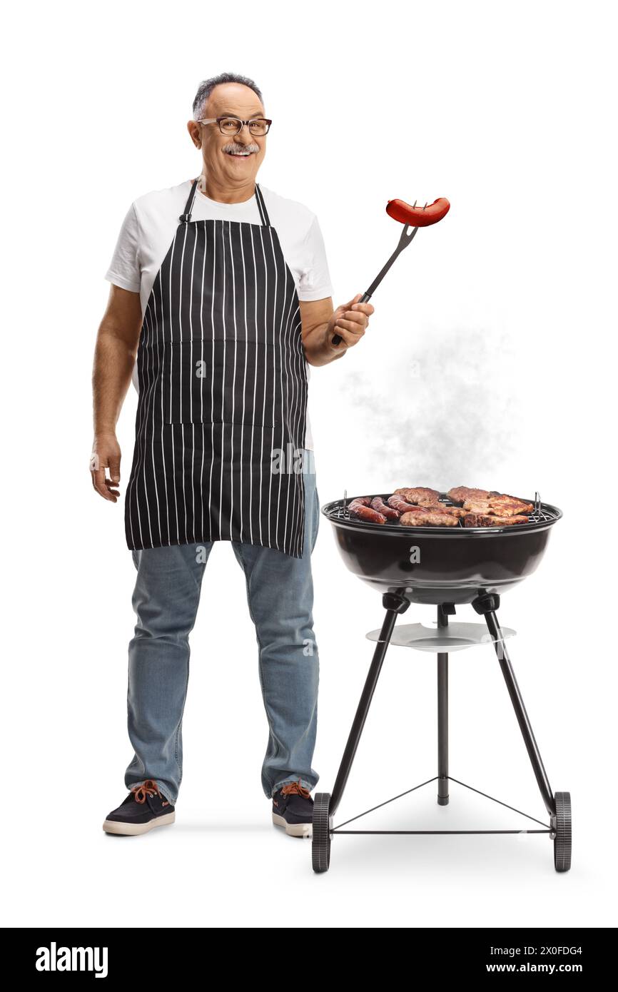 Uomo maturo accanto a un barbecue portatile che tiene in mano una salsiccia con una forchetta isolata su sfondo bianco Foto Stock